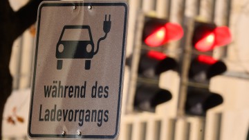 Ein Schild mit einem Symbol für ein Elektroauto und den Schriftzug „während des Ladevorgangs“ markiert einen Parkplatz an einer Ladesäule. Im Hintergrund leuchten die drei Farben einer Ampel auf. Nach dem plötzlichen Ende der Kaufprämie für Elektroautos s