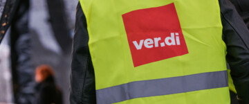 Detailaufnahme: Ein Beschäftigter mit einer Warnweste, auf der ein Verdi-Logo abgebildet ist, steht im Vordergrund (Symbolfoto)