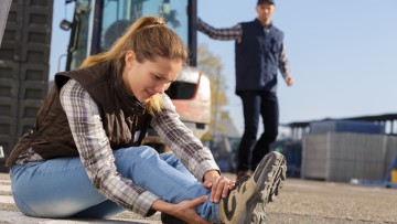 Eine junge Frau ist auf einem Betriebshof gestürzt und hält sich den Knöchel. Im Hintergrund ein Gabelstaplerfahrer, der zu Hilfe eilt. (Symbolbild mit Fotomodellen)