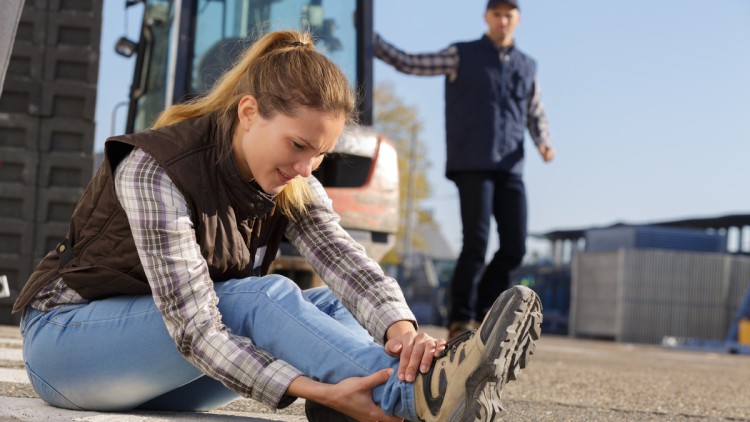 Eine junge Frau ist auf einem Betriebshof gestürzt und hält sich den Knöchel. Im Hintergrund ein Gabelstaplerfahrer, der zu Hilfe eilt. (Symbolbild mit Fotomodellen)