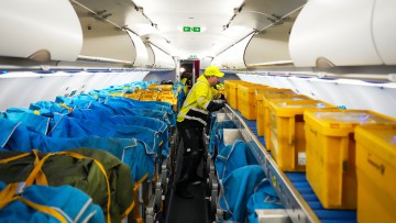 Ein Mitarbeiter der WISAG schiebt in einem Airbus A320-214 gelbe Plastikboxen voller Briefe und anderer Post in den vorderen Teil der Kabine. Die Passagiersitze wurden zuvor mit Stoffmanschetten abgedeckt, in denen die Postboxen mit Gurten festgezurrt sin