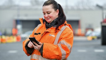Frau in orangener BLG Logistics-Jacke schaut fröhlich auf ihr Handy