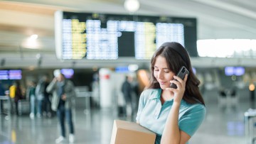 Frau am Flughafen mit Paket in der Hand und Handy am Ohr