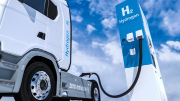 Lkw lädt an einer Tankstellen-Zapfsäule mit Wasserstoff-Logo vor weiß-blauem Himmel