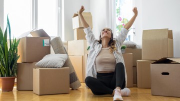 Junge Frau sitzt mit Kisten auf dem Boden in neuer Wohnung und hebt freudig die Arme