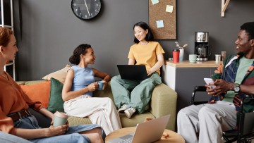 Eine Gruppe junger Leute sitzt entspannt im Büro neben einer Kaffeemaschine mit Laptops und reden miteinander
