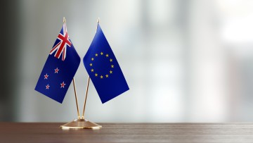 Zwei kleine Flaggen von Neuseeland und der EU stecken in einer goldenen Halterung auf einem leeren Holzschreibtisch