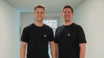Philipp Henn und Roland Moussavi lächeln in schwarzen T-Shirts in die Kamera