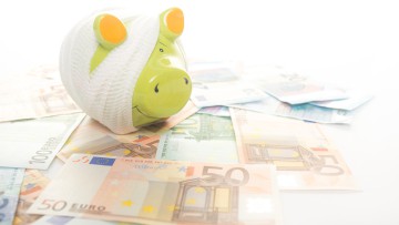 Ein „krankes“ Sparschwein – mit einem Verband umwickelt – steht auf Geldscheinen. Symbolbild für Krankengeld