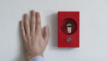 Eine Hand liegt neben einer Art roten Alarmkasten, in der eine Trillerpfeife (englisch: Whistle) durch ein rundes Sichtloch aufgehängt zu erkennen ist