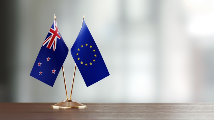 Zwei kleine Flaggen von Neuseeland und der EU stecken in einer goldenen Halterung auf einem leeren Holzschreibtisch