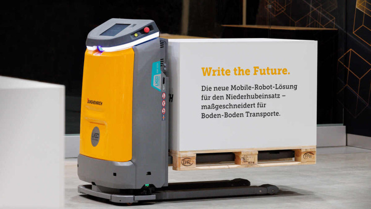 Der Autonomous Mobile Robot (AMR) EAE 212a von Jungheinrich transportiert auf einer Europalette eine Box mit der Aufschrift "Write the Future. Die neue Mobile-Robot-Lösung für den Niederhubeinsatz - maßgeschneidert für Boden-Boden-Transporte."