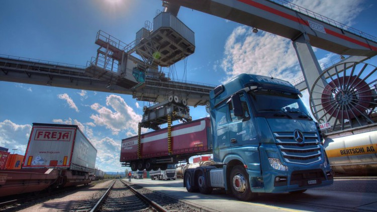 In einem Umschlagsterminal der DB Cargo hat eine Maschine einen Trailer hinter einer Zugmaschine aufgenommen, um ihn auf einen Güterwagen abzusetzen. Rechts und Links davon stehen Gleise mit  Güterzügen